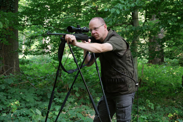 Jäger mit Schießstock im Wald, dabei die Savane-Weste tragend, braune Seite außen