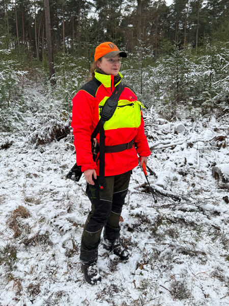 Jägerin bei trübem Wetter in verschneitem Brachland vor einem Waldstück stehend, dabei Hundeführerjacke und Sauenschutzhose tragend und visuell auffällig aus der weiß-grauen Landschaft hervorstechend