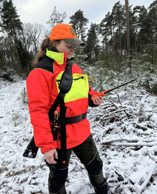 Jägerin bei trübem Wetter in verschneitem Brachland vor einem Waldstück stehend, dabei die PSS-Jacke tragend und visuell auffällig aus der weiß-grauen Landschaft hervorstechend